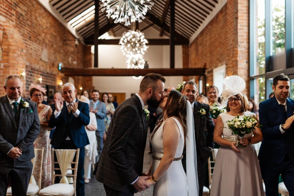 Elegant wedding ceremony at a charming historic venue with exposed brick walls, wooden beams, and modern lighting, featuring a kissing couple surrounded by joyful guests.