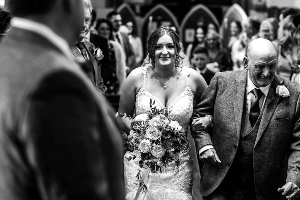 Elegant bride walking down the aisle in a wedding ceremony, holding a floral bouquet, surrounded by family and friends, captured in black and white for timeless wedding memories.