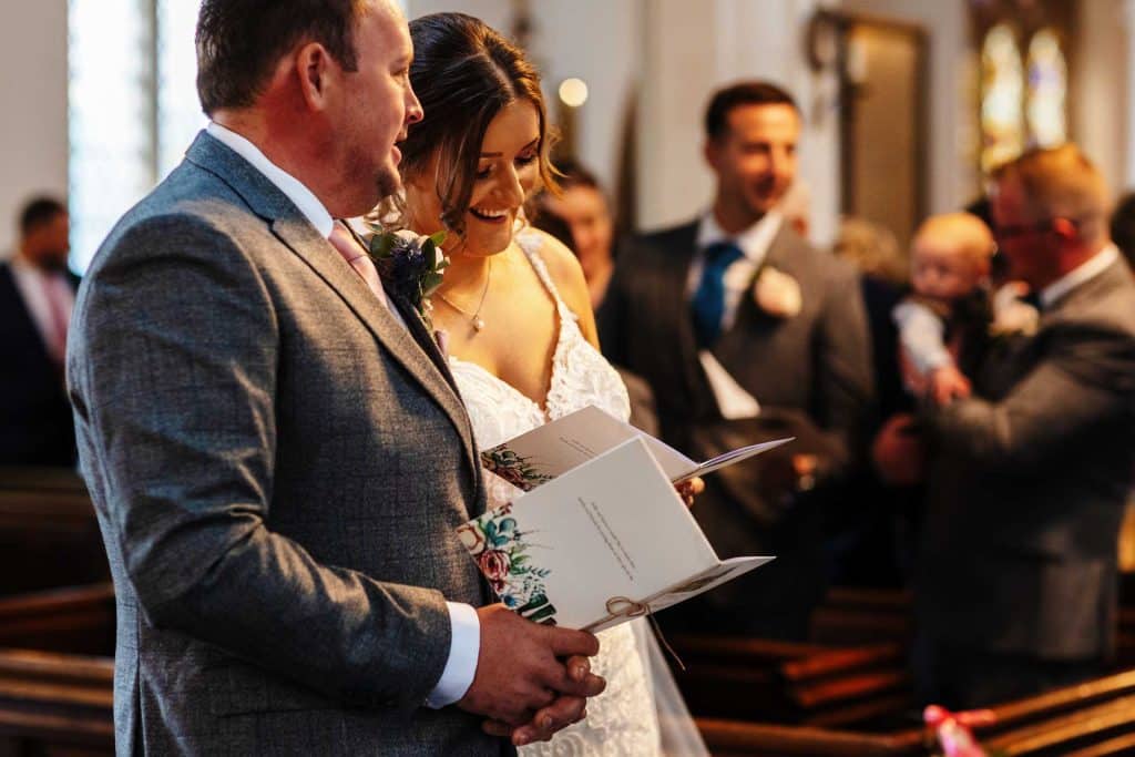 Elegant wedding ceremony with bride and groom reading vows in church, surrounded by wedding guests, capturing joyful matrimonial moments for BGS Weddings.