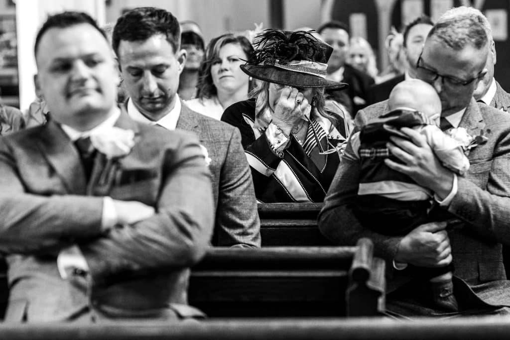 Black and white photo of wedding guests inside a church, capturing emotional moments, with a woman in a hat wiping tears, emphasising the significance of the wedding ceremony.