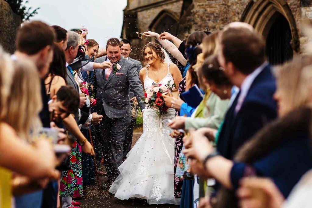 Elegant bride and groom celebrate wedding surrounded by friends and family at BGS Weddings, capturing joyful moments outside a historic venue with confetti and floral arrangements.