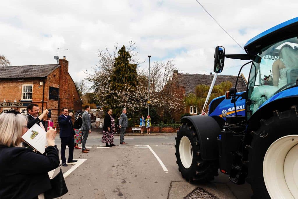 Groom and wedding guests taking photos of bride arriving on tractor at rural wedding in England; wedding transportation, countryside nuptials, outdoor wedding celebration, bride’s arrival, BGS Weddings.