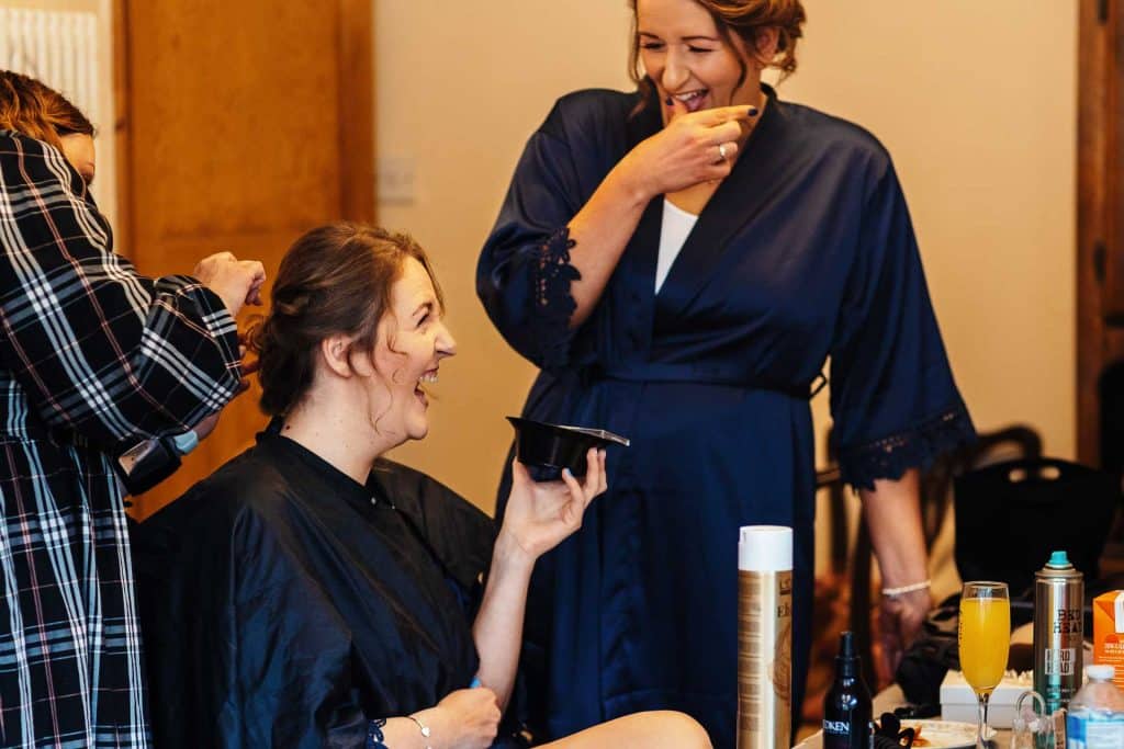 Uneven makeup application for a bride before her wedding, with her sister helping her and friends preparing in the background, showcasing professional wedding hair and makeup services by BGS Weddings.