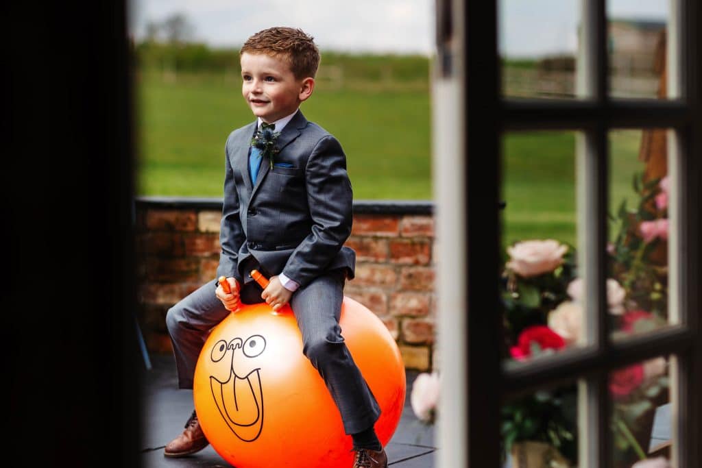 Bright young boy in a grey suit riding an orange balloon with a silly face on a wedding venue, capturing a playful wedding moment, outdoor celebration, children’s entertainment at BGS Weddings.