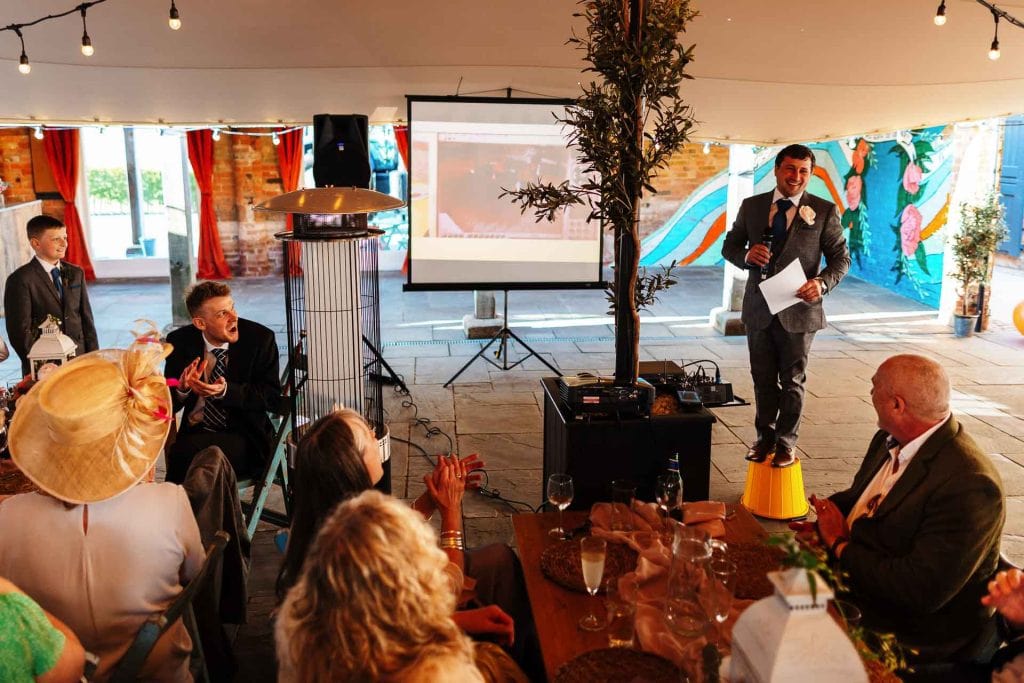 Multiple wedding guests enjoying a lively speech at a rustic indoor wedding reception with colourful decorations and a relaxed atmosphere.