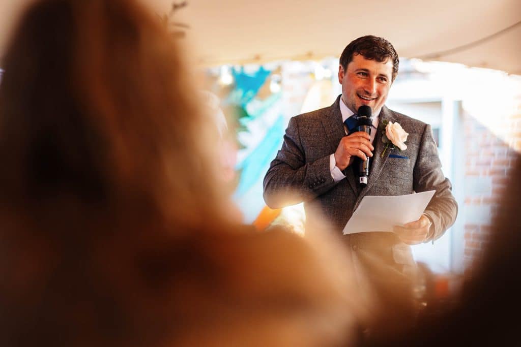 Warmly dressed groom giving speech at wedding reception, holding a microphone and notes, smiling during his heartfelt toast.