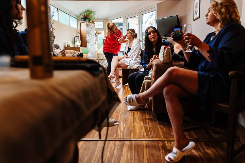 Applying makeup for the bride in a cosy, well-lit living room before her wedding at BGS Weddings, with bridesmaids and friends preparing and relaxing together for a memorable special day.