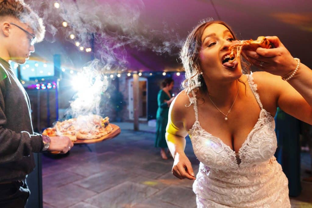 Delighted bride enjoying a slice of pizza at her wedding reception, capturing a joyful moment during the celebration.