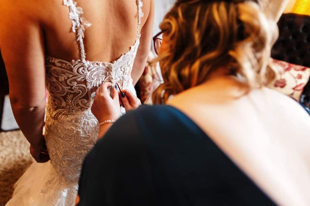 Elegant bride getting ready for wedding, lace wedding dress being fastened by a helper at BGS Weddings.
