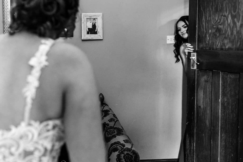 Bride peeking from behind a door, preparing for her wedding, with wedding decor and family photos on the wall, captured in black and white.