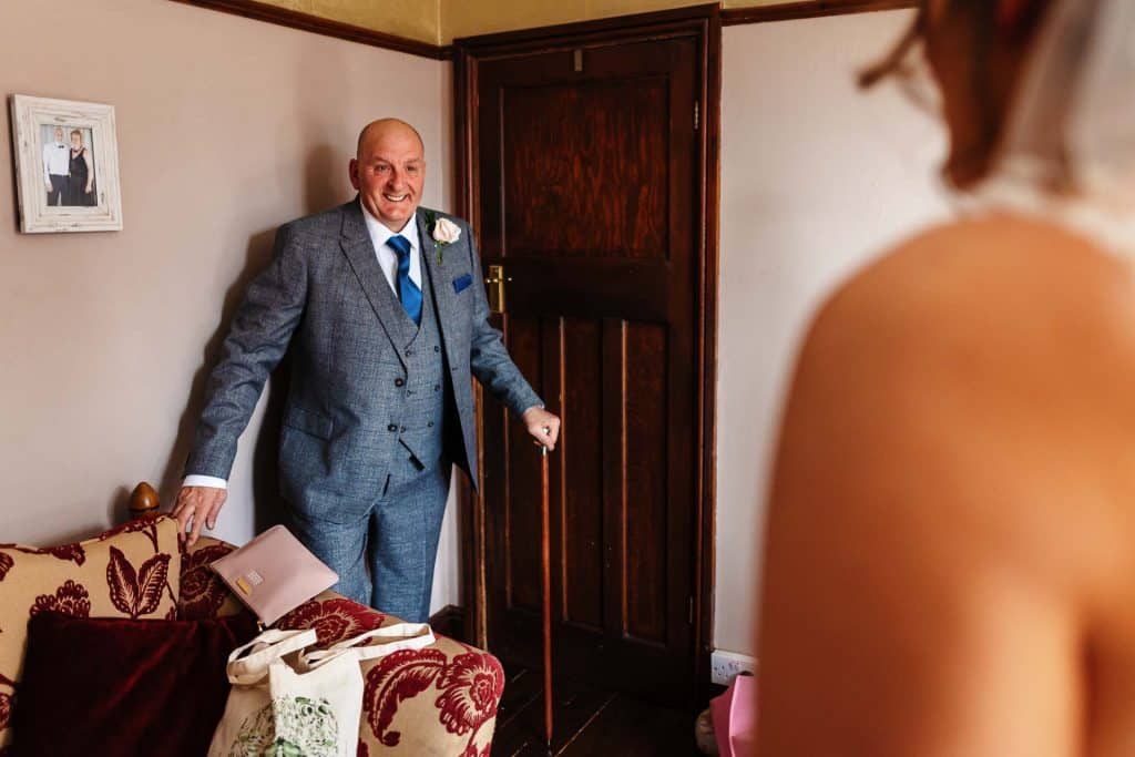 Happy groom in grey suit with blue tie and boutonnière, standing at house door before wedding, surrounded by wedding accessories, captured in a warm, intimate moment.