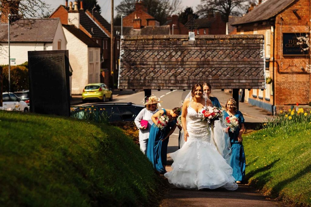 Elegant bride in a white wedding gown walking with bridesmaids and flower girl along a scenic village street, capturing memorable wedding moments at BGS Weddings.