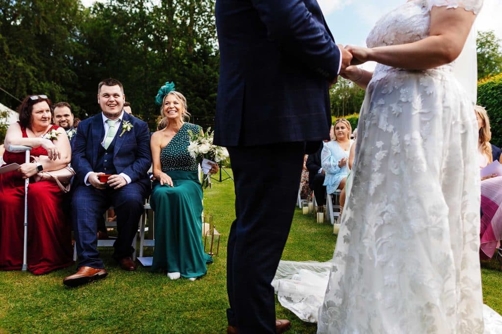 Elegant outdoor wedding ceremony with the bride and groom exchanging vows, surrounded by cheerful guests, lush greenery, and floral details, captured by BGS Weddings in the UK.