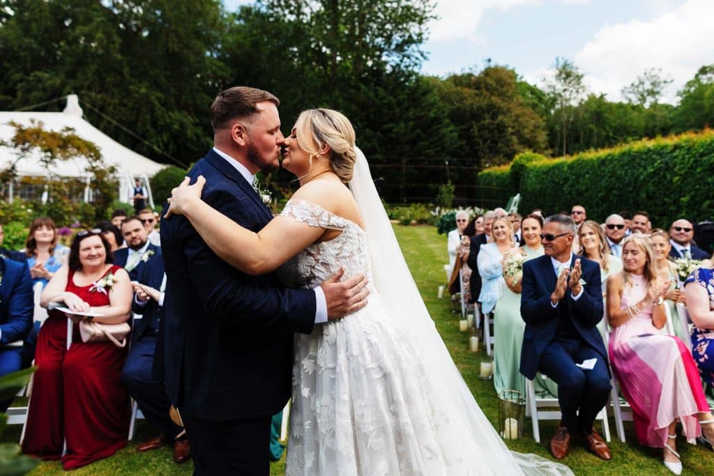 Beautiful wedding couple sharing a kiss during outdoor wedding ceremony at BGS Weddings venue. Elegant bride in lace gown and groom in navy suit celebrate love surrounded by cheering guests.
