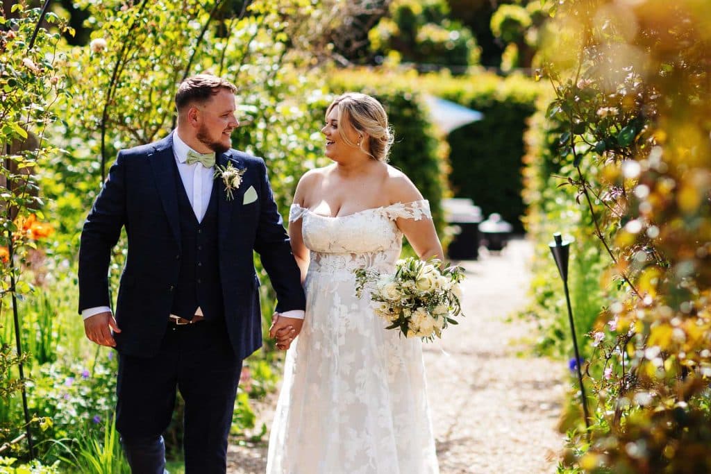 Elegant wedding couple walking hand in hand through a lush garden at BGS Weddings, showcasing beautiful outdoor wedding photography in a stunning floral setting.