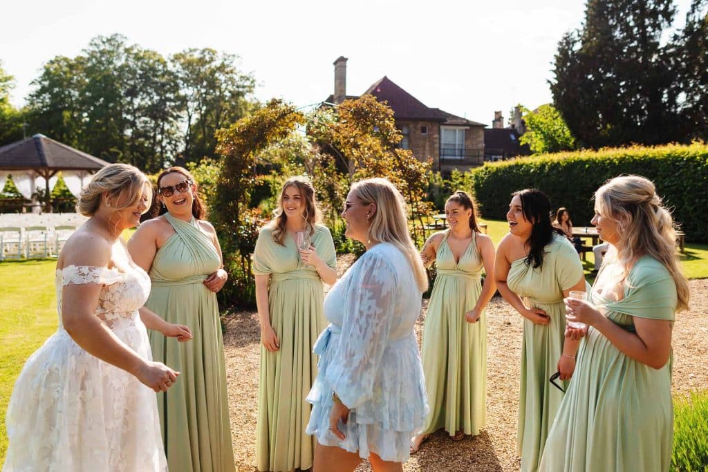 Elegant bride and bridesmaids celebrating outdoors at a wedding, showcasing bridal gown and pastel dresses, with lush garden setting, bright sunlight, and joyful atmosphere at BGS Weddings UK.