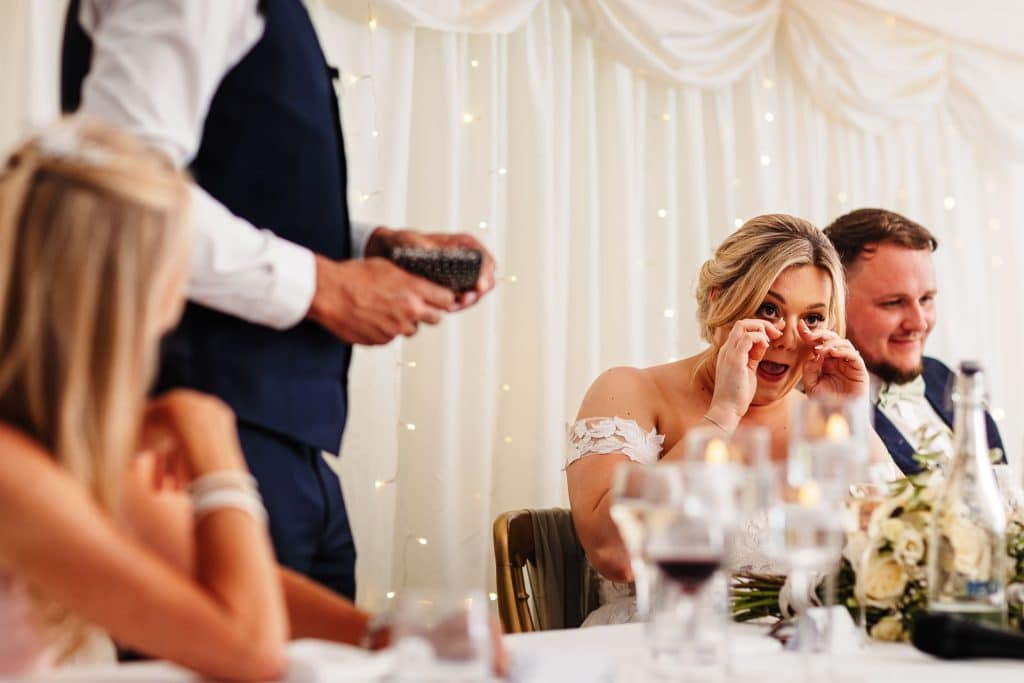 Beautiful bride wiping tears of joy at wedding reception, surrounded by loved ones, elegant decor, and fairy lights, captured by BGS Weddings for memorable wedding photography.