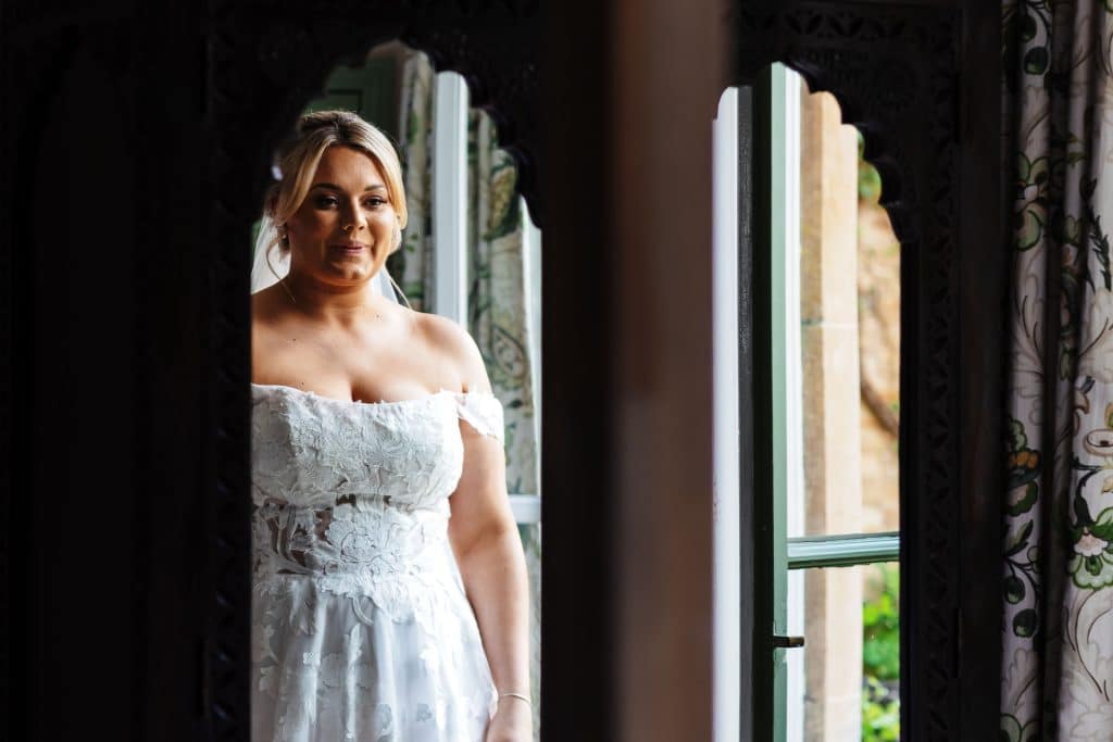 Bride preparing for her wedding day at BGS Weddings, looking out the window in her elegant off-shoulder lace wedding dress, captured in intimate detail.