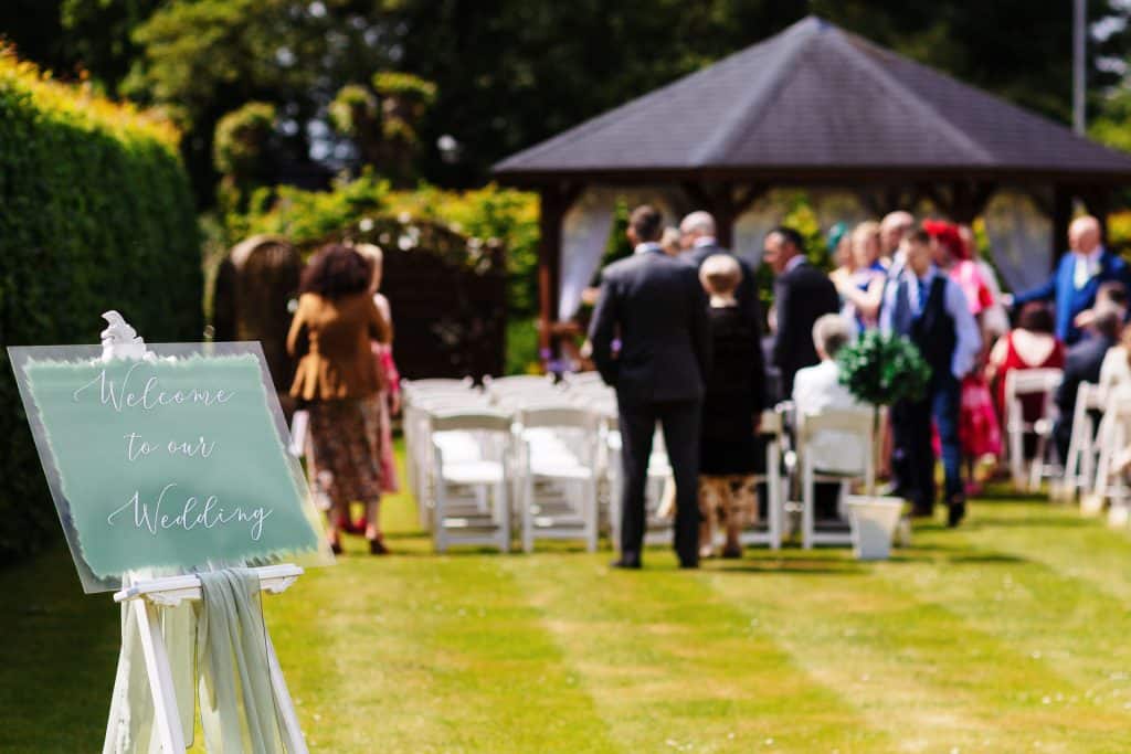Elegant outdoor wedding ceremony with guests, lush garden setting, and welcoming sign at BGS Weddings in the UK.