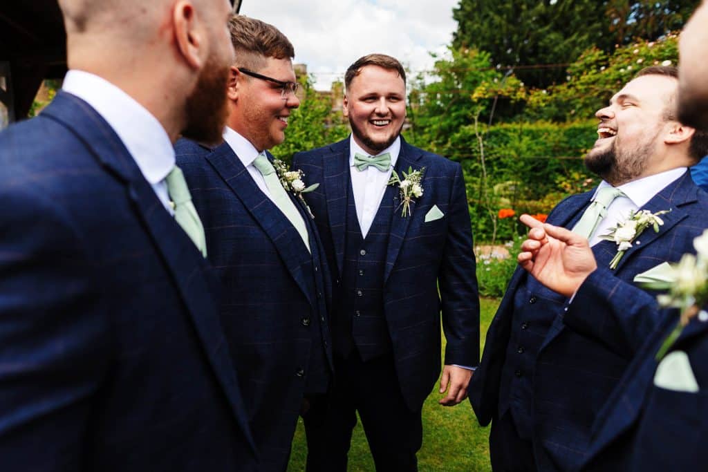 Group of groomsmen in matching navy suits with light green ties and boutonnières, chatting and sharing laughs outdoors during a wedding celebration.