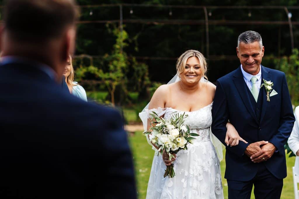 Elegant bride in a white lace wedding dress holding a bouquet smiles as she walks down the aisle with her father at an outdoor wedding ceremony in a lush garden setting.