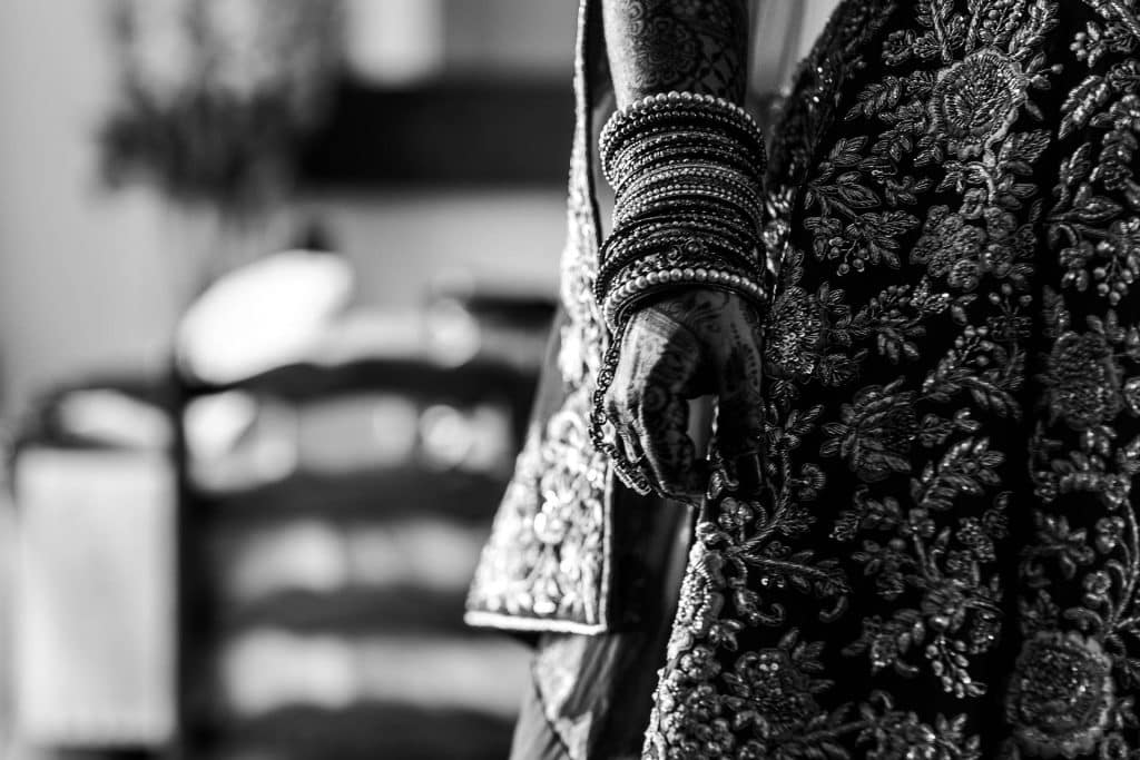 Intricate close-up of a bride’s hand and wrist adorned with traditional bangles and henna, highlighting the detailed embroidery of her bridal outfit at a wedding.