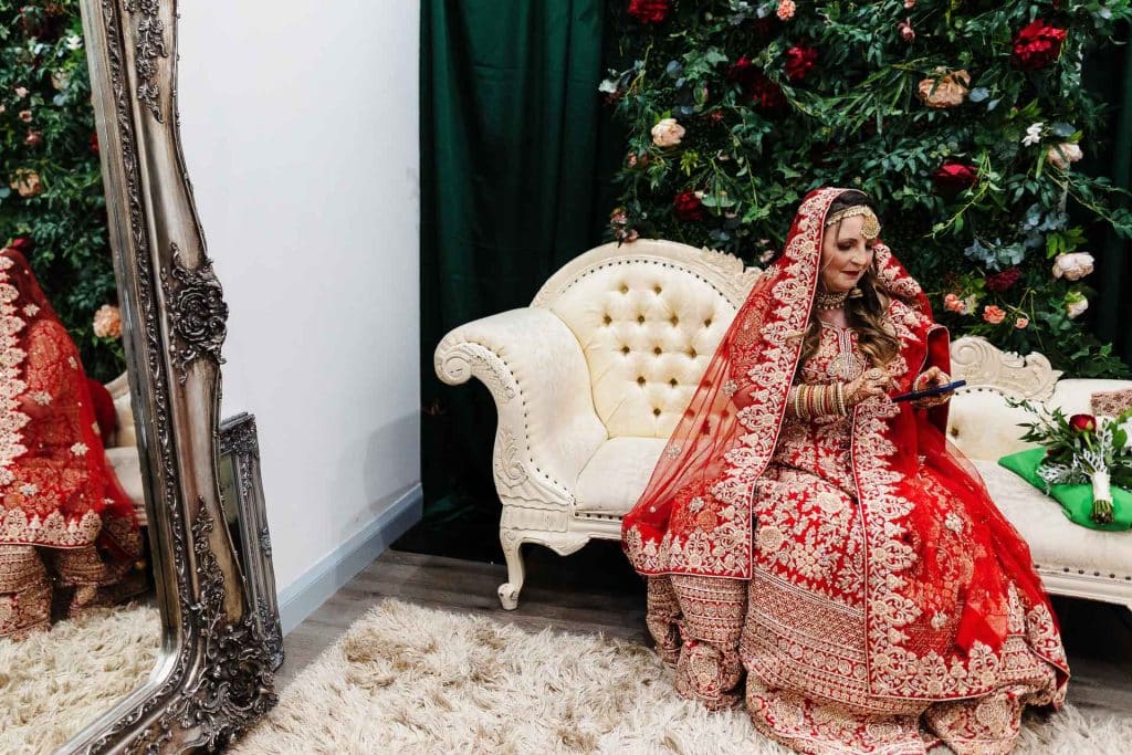 Elegant bride in traditional red wedding attire sitting on a cream vintage sofa, surrounded by floral backdrop and ornate mirror, capturing the essence of a luxury Indian or South Asian wedding.