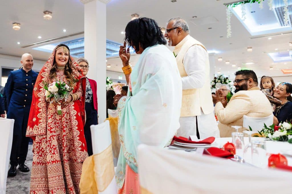 Elegant bride in traditional red bridal attire holding flowers at multicultural wedding reception.