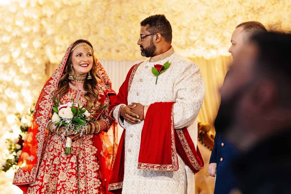 Vibrant Indian wedding ceremony with bride and groom in traditional attire exchanging vows under elegant floral décor. Celebrating love and cultural traditions at BGS Weddings UK.