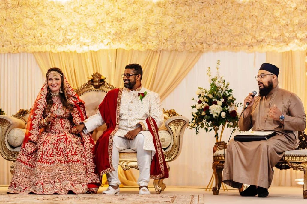 Elegant South Asian wedding ceremony with bride and groom seated on ornate throne-style chairs, traditional attire, floral decor, and an officiant speaking with a microphone.