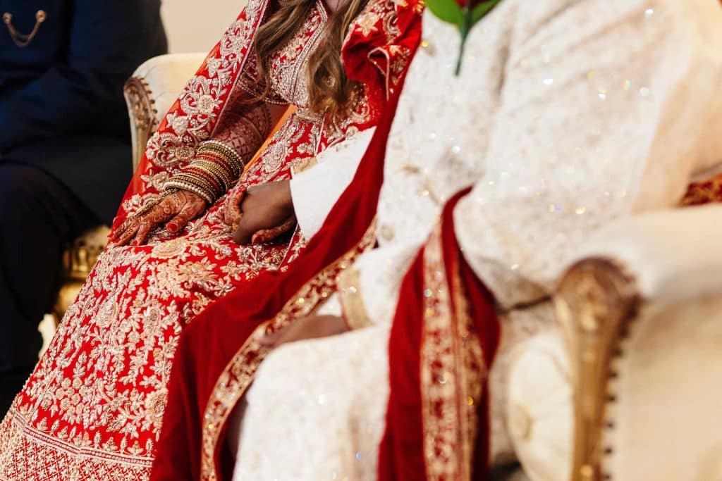 Elegant Indian wedding brides seated on ornate chairs showcasing traditional red and white bridal attire with intricate embroidery and gold jewelry for a cultural wedding celebration.