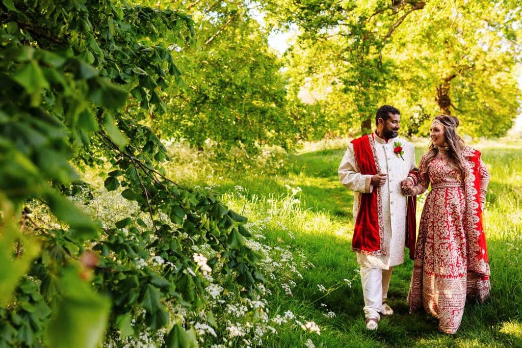 Elegant Indian wedding couple walking in a lush green park with vibrant traditional attire, surrounded by trees and blooming flowers, capturing a joyful moment of multicultural wedding celebration.