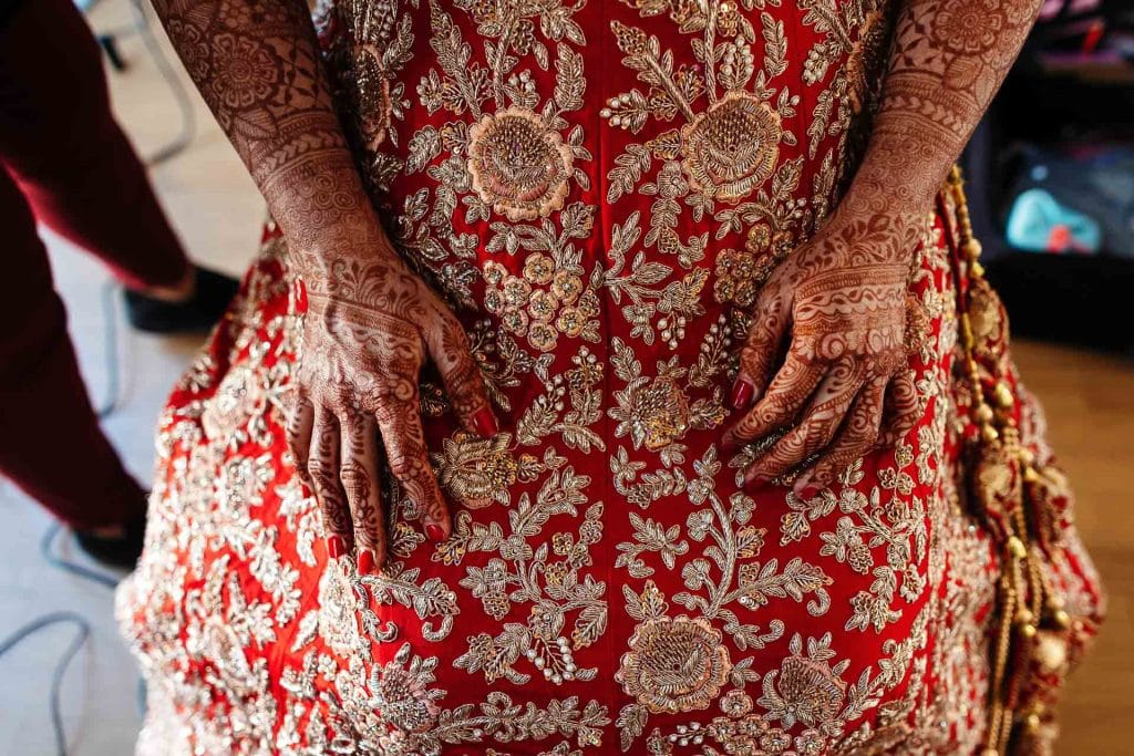 Intricate henna designs on hands and traditional red and gold bridal attire, showcasing detailed embroidery and cultural wedding elements for an Indian wedding.