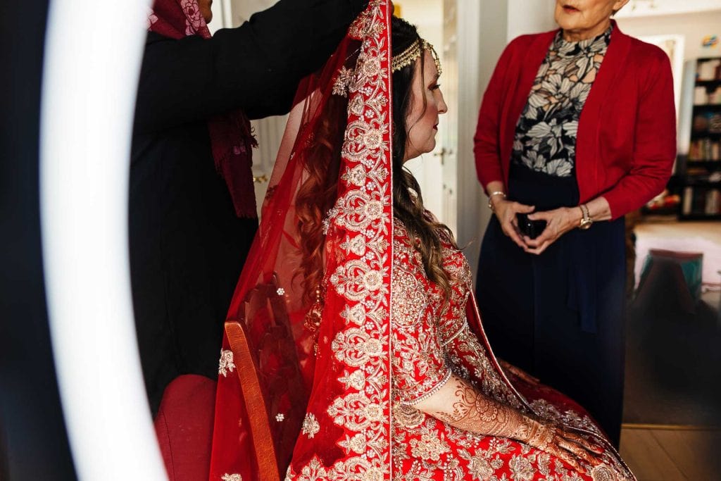 Elegant bride in intricate red and gold wedding attire with henna designs, surrounded by family members preparing for wedding ceremony at BGS Weddings UK.