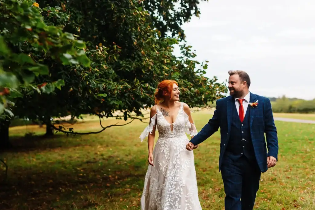 Elegant wedding couple walking hand in hand outdoors, bride in a beautiful lace wedding dress and groom in a tailored check suit with a red tie, surrounded by lush greenery, outdoor wedding, BGS Weddings.