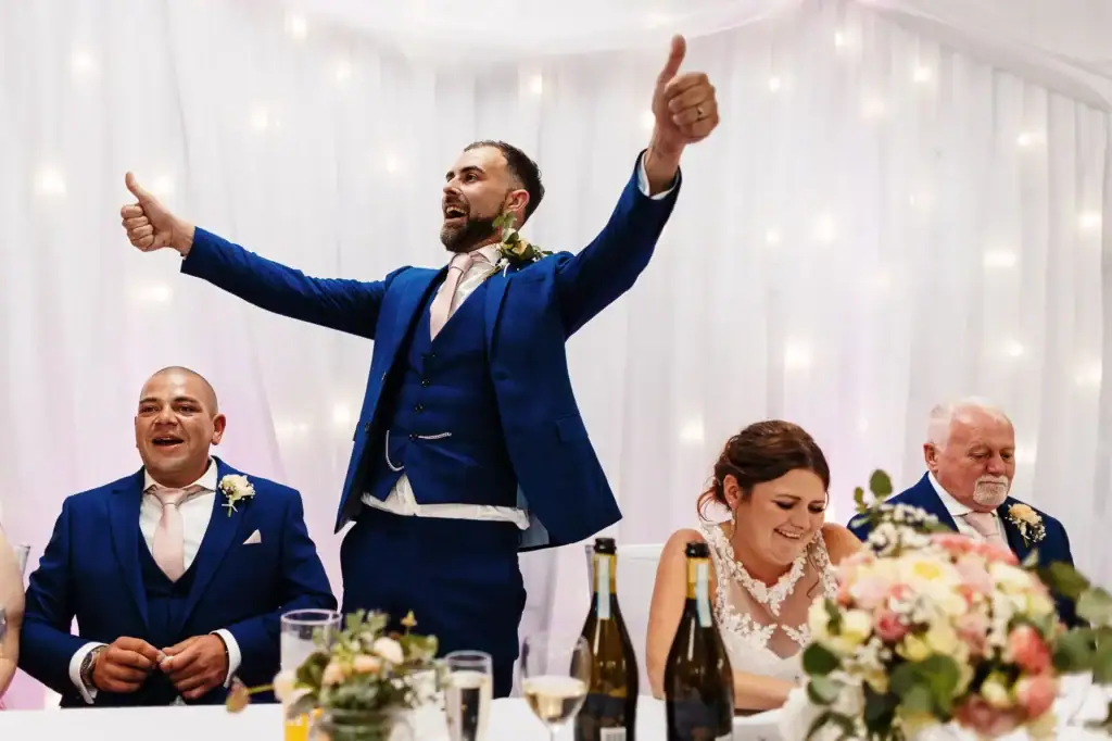 Celebration of wedding speeches with joyful groom giving a toast at a wedding reception with happy guests, elegant decor, and floral arrangements.