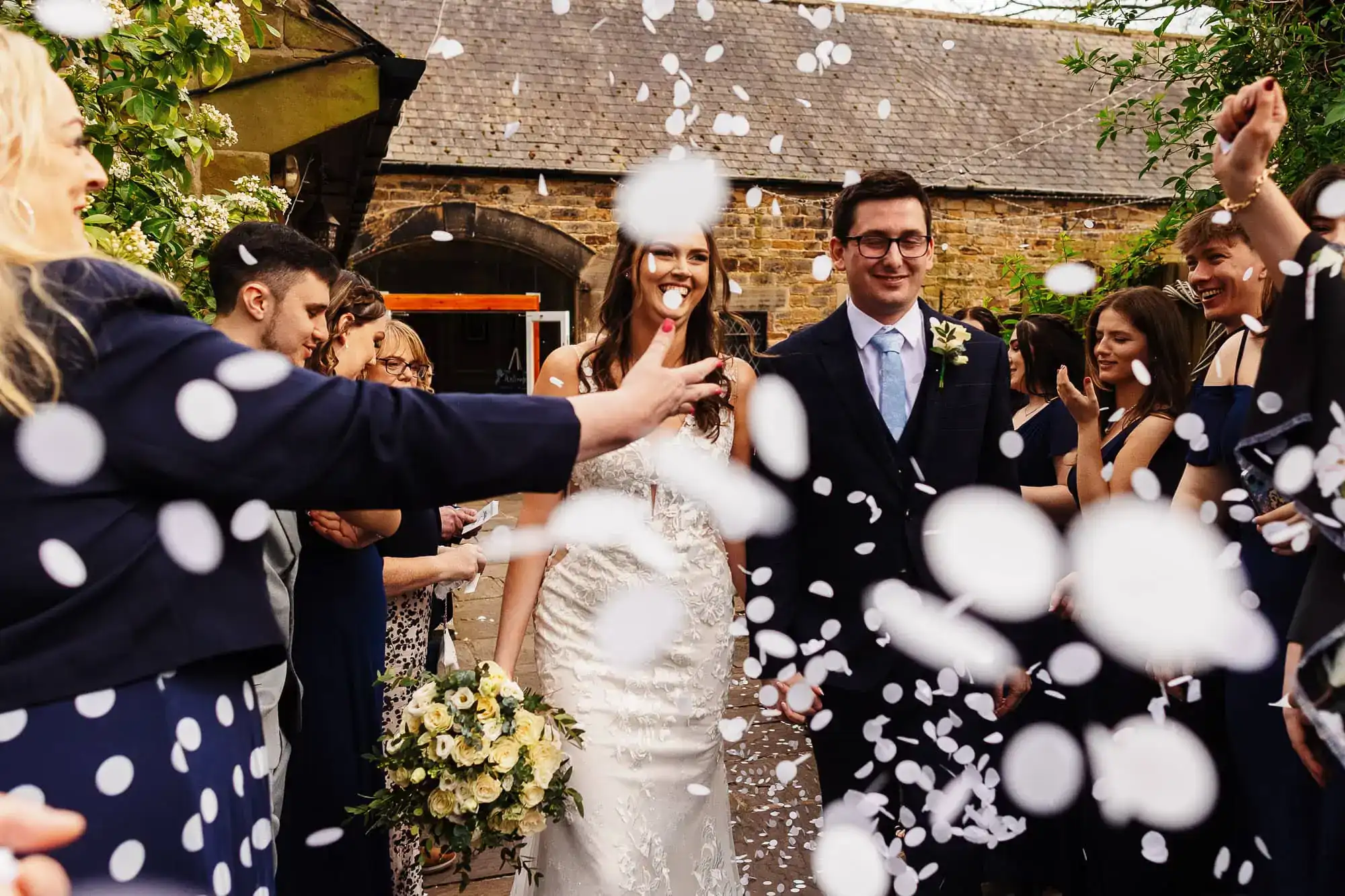 Elegant bride and groom celebrating wedding ceremony surrounded by friends and family with confetti outdoor at historic venue.