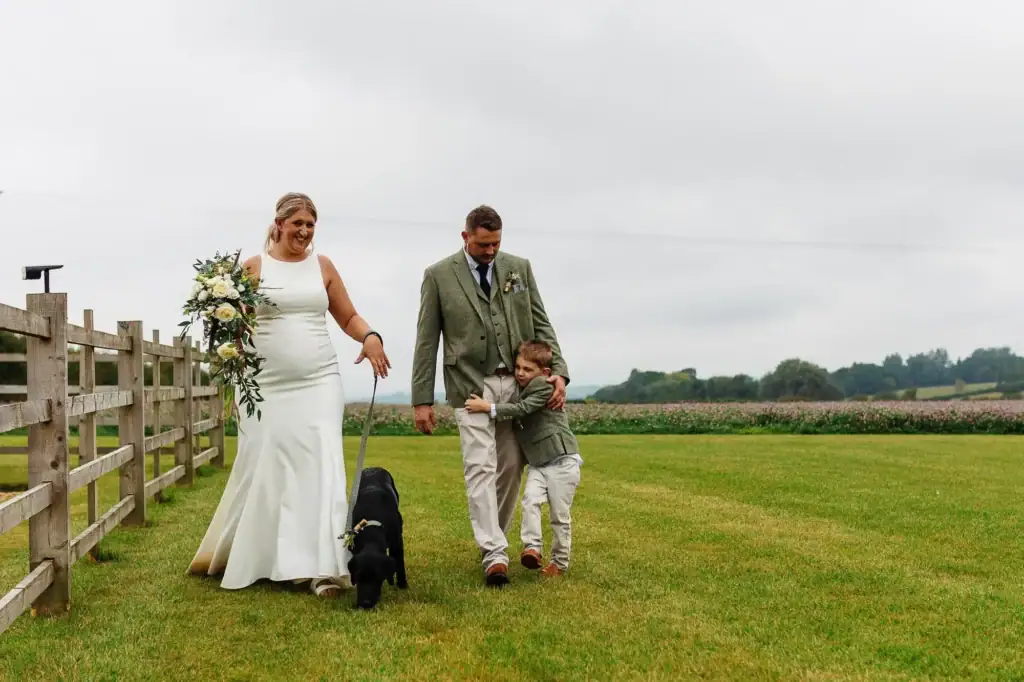 Elegant bride and groom with a young boy and black Labrador dog walking in a scenic outdoor wedding setting, captured by BGS Weddings, showcasing natural and memorable wedding moments.
