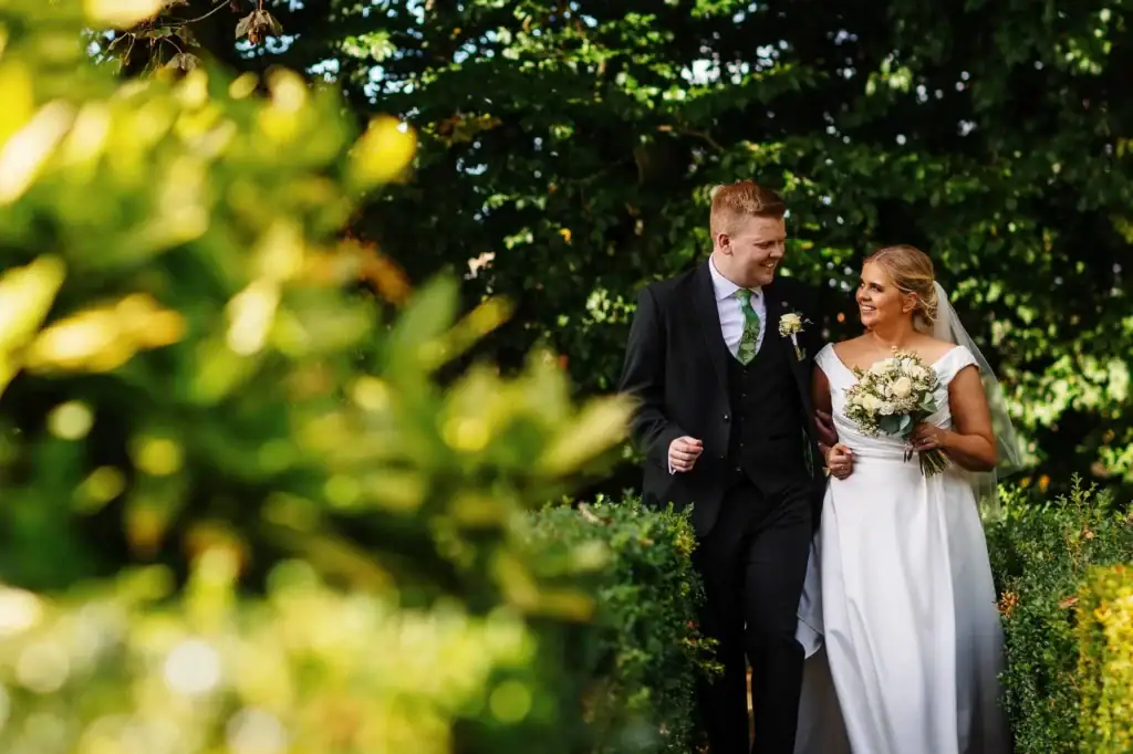 Wedding couple walking through lush garden, bride in white gown holding bouquet, groom in black suit with floral tie, romantic outdoor wedding, BGS Weddings UK, elegant wedding photography, picturesque wedding shot in green setting.