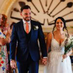 Elegant wedding couple holding hands after ceremony in a beautifully decorated venue, surrounded by happy guests celebrating a memorable wedding day.