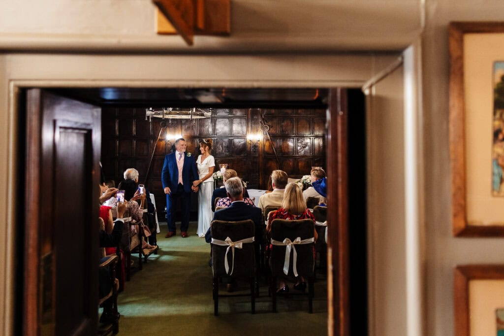Elegant wedding ceremony with bride and groom holding hands, surrounded by family and friends, captured through a doorway at a charming UK wedding venue.