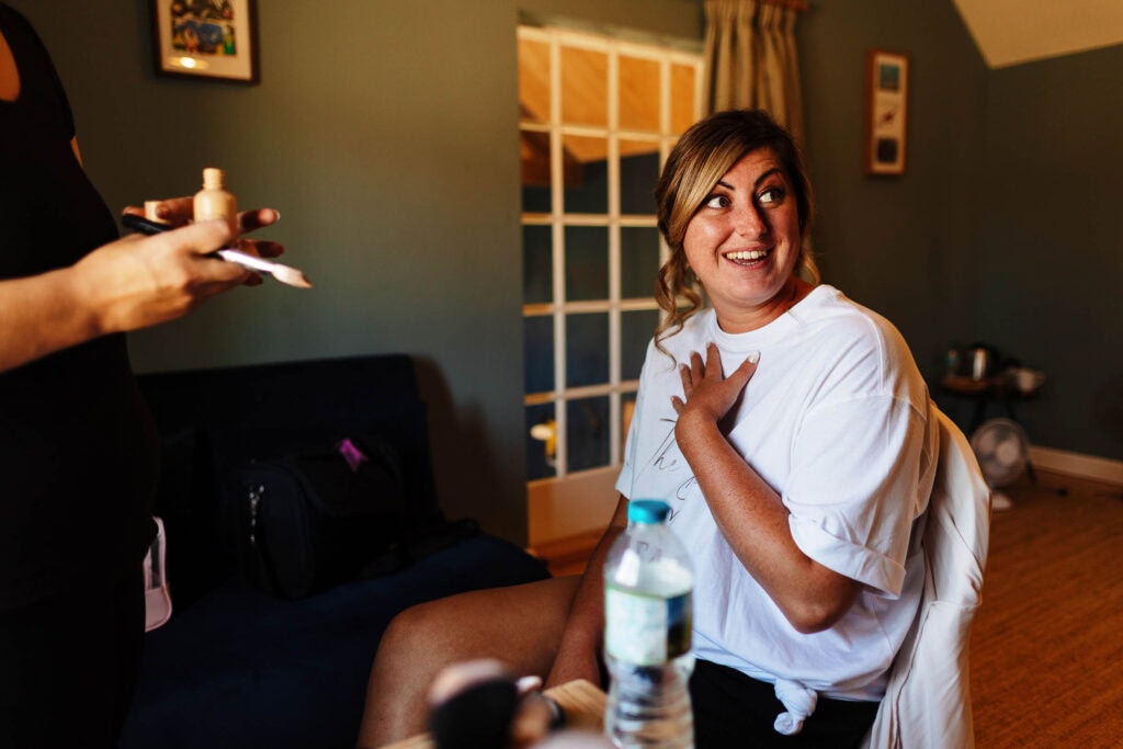 Bride getting ready for her wedding at BGS Weddings, natural light, joyful moment, makeup artist applying finishing touches, indoor setting with stylish decor, wedding preparation scene.
