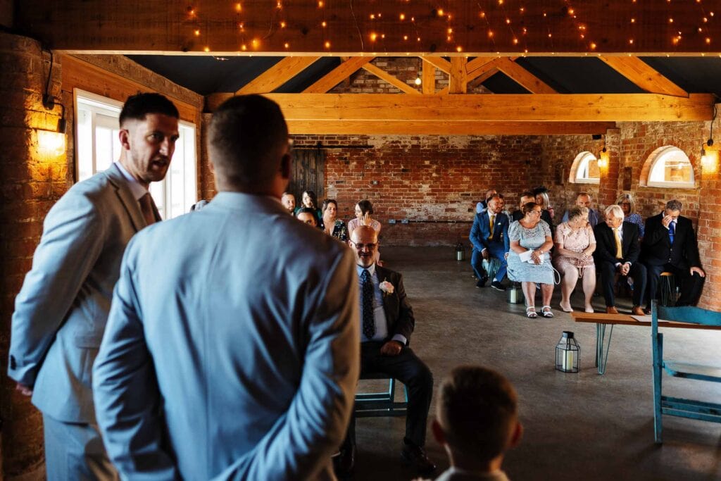Elegant wedding ceremony in rustic barn venue with guests seated, wooden beams, and string lights, captured by BGS Weddings.