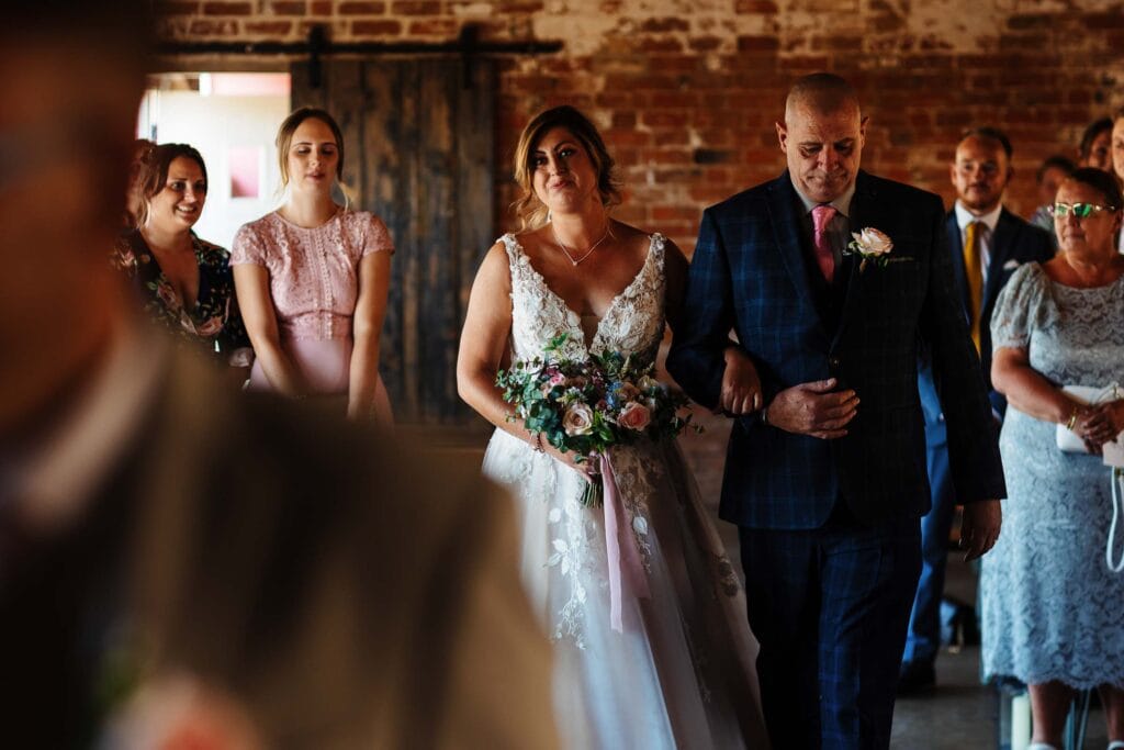 Elegant bridal procession at a rustic wedding venue with guests, bride in lace dress holding bouquet, escorted by father in checkered suit, capturing memorable wedding moments.