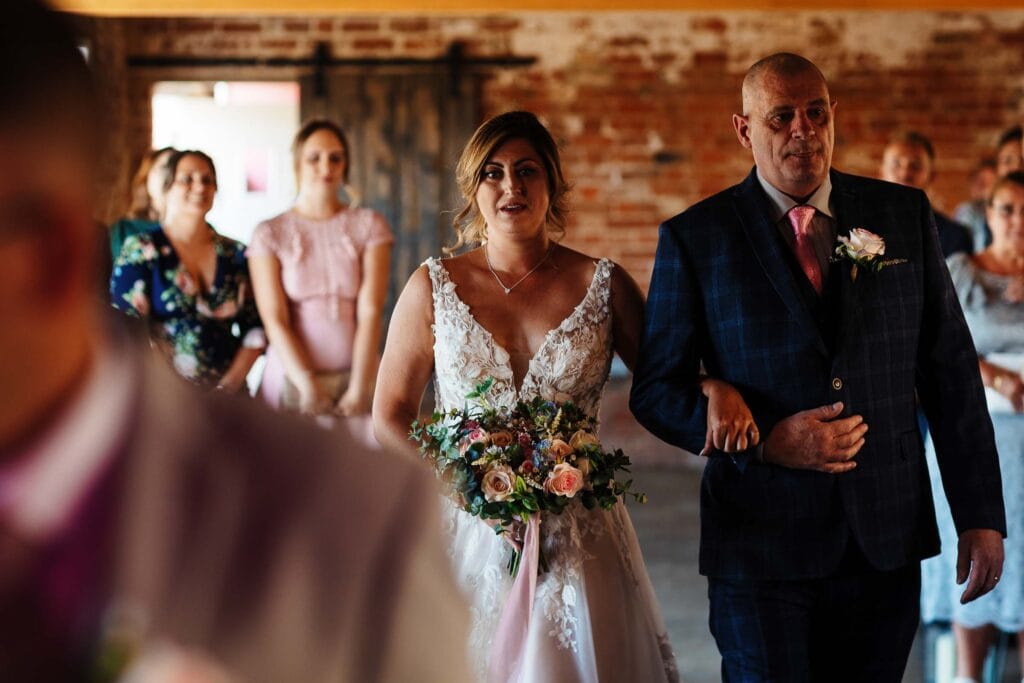 Elegant bride holding a floral bouquet walking down the aisle during a rustic wedding ceremony at BGS Weddings, with her father or relative.
