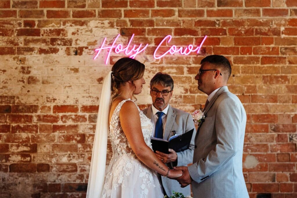 Elegant wedding ceremony at Holy Cow venue featuring a bride and groom exchanging vows in front of a brick wall with neon pink "Holy Cow" sign, highlighting modern wedding styles in the UK.
