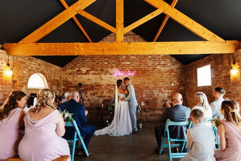 Romantic wedding ceremony at BGS Weddings with couple sharing a kiss in an indoor rustic venue with exposed brick walls and wooden beams.