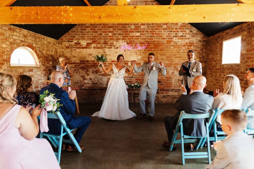 Joyful wedding celebration with bride and groom raising hands in rustic brick venue decorated with fairy lights and neon sign.