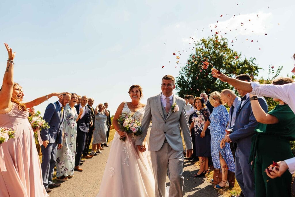 Elegant wedding couple walking hand in hand with joyful guests throwing confetti, outdoor wedding celebration, beautiful bride and groom, romantic ceremony, wedding day happiness, BGS Weddings UK.