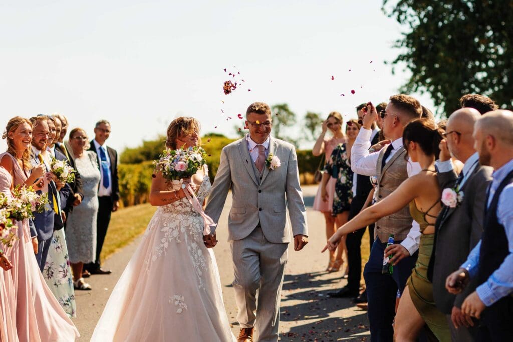 Beautiful wedding celebration with newlyweds walking hand in hand surrounded by joyful guests, rose petal toss, outdoor wedding, elegant bridal gown, stylish groom, wedding party, BGS Weddings.